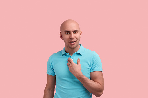 Why me. Young bald homosexual man with bristle indicates on himself with hand, fights for equity and rights, argues, wants know truth, gay friendly, wears blue polo shirt, poses over pink background