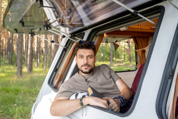 istock Young bearded man with book looking at camera in window 1339665904