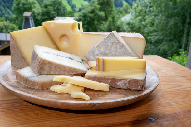 käsesammlung, holzbrett mit französischen käsesorten comte, beaufort, abondance, emmental, morbier und französisches bergdorf in haute-savoie im hintergrund - morbier stock-fotos und bilder