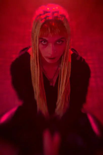 Young woman on her knees with devilish look in her eyes, red lights, horror concept