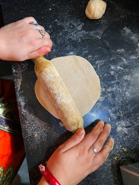 Close-up image of unrecognisable person preparing a roti / chapatti with rolling pin on kitchen counter, wholewheat atta flour dough rolled into disk, elevated view, focus on foreground stock photo