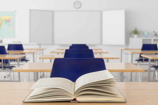 Open Book On Wooden Desk With Blurred Empty Classroom Background.