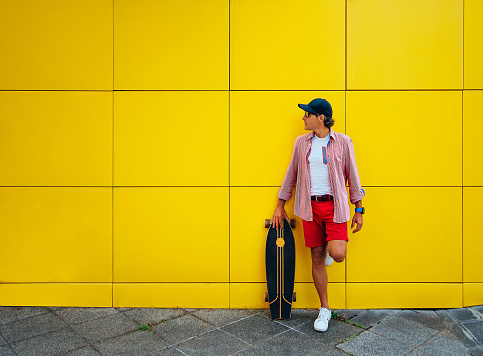 middle-aged man with a cap, leaning against a yellow wall with a skateboard.the person looks to the side