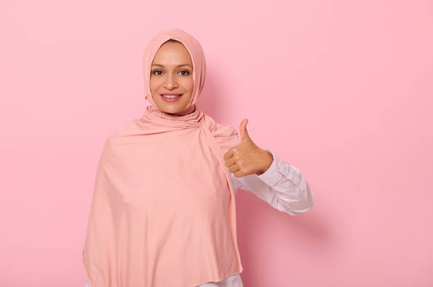 adorable et charmante femme arabe en hijab montre un pouce levé et sourit avec un beau sourire denté en regardant la caméra, isolée sur fond rose avec espace de copie - confidence toothy smile thumbs up ok sign photos et images de collection