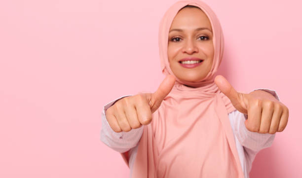 concentrez-vous sur les mains d’une femme arabe musulmane au sourire flou avec un sourire édenté en hijab rose, montrant les pouces vers la caméra, isolés sur un fond coloré avec de l’espace pour le texte - confidence toothy smile thumbs up ok sign photos et images de collection
