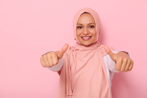 une femme arabe musulmane amicale et charmante en hijab rose montre les pouces levés à la caméra, sourit avec un sourire denté, isolée sur fond coloré avec un espace de copie - confidence toothy smile thumbs up ok sign photos et images de collection