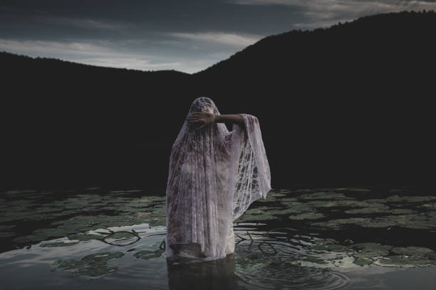 Bride in a white wedding dress standing in a lake surrounded by water lilies, covering her face, night is coming. Mysterious ghost like woman stares at the camera as night approaches. Halloween horror concept. scary bride stock pictures, royalty-free photos & images