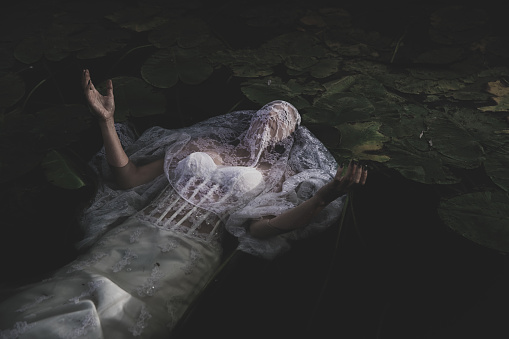 Woman in a white wedding dress looking like Opehlia lies in a dark pond, holding hands up, praying.