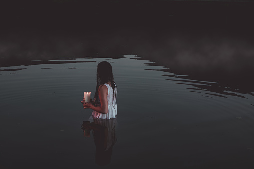 The ghost of a woman in a white dress holds a white candle and stands in a pond. Halloween horror concept.