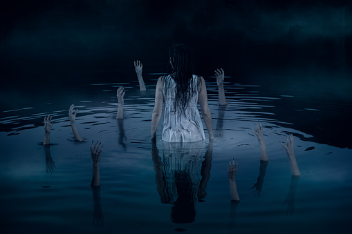 Hands sticking out of the water, reaching out to the woman in white night dress. Halloween horror concept.