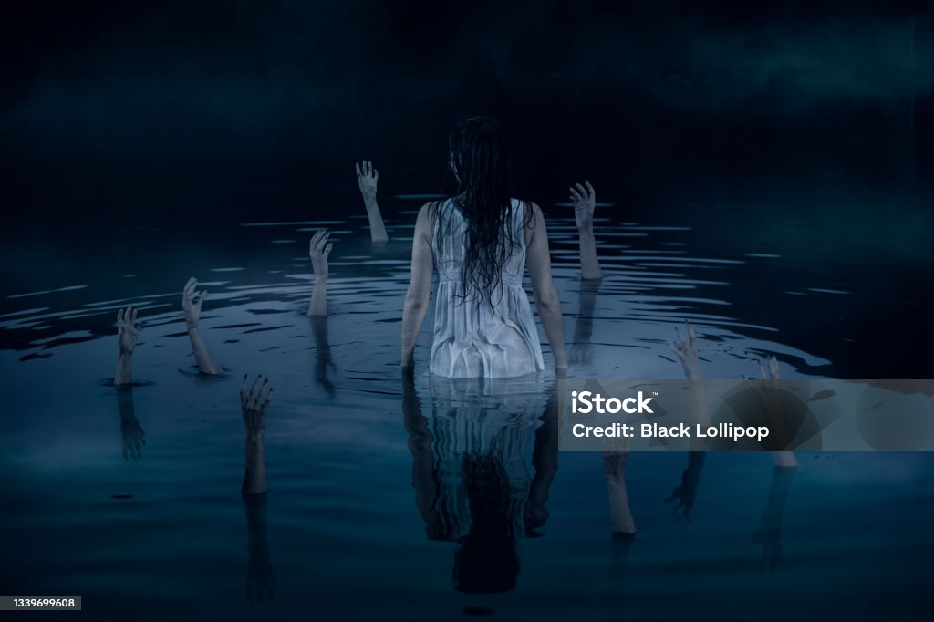 A woman stands in the middle of the lake surrounded by arms reaching out of deep dark water. Hands sticking out of the water, reaching out to the woman in white night dress. Halloween horror concept. Horror Movie Stock Photo