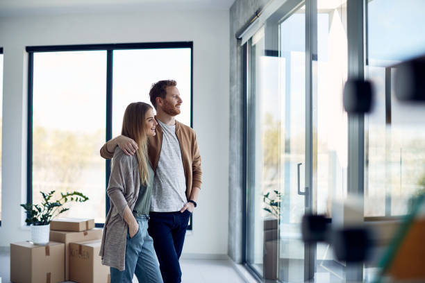 jeune couple embrassé regardant par la fenêtre de leur nouvel appartement. - location immobilière photos et images de collection