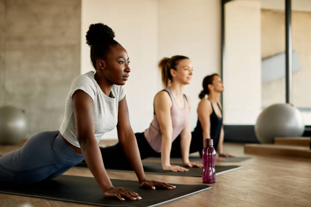 Black female athlete doing stretching exercises while warming up with group of women at health club. Athletic women doing stretching wile exercising on group training in a gym. Focus is on African American woman. yoga class stock pictures, royalty-free photos & images