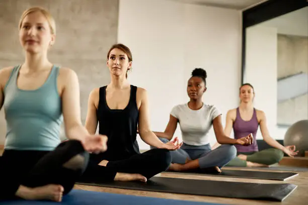 Young athletic women meditating in lotus pose during Yoga class at health club. Focus is on woman in black.