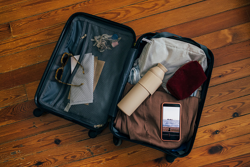 A packed suitcase from above on the wooden floor on a holiday or a business trip containing a water bottle, an unlocked smartphone, a cosmetic bag, sunglasses, plane tickets, clothes and keys