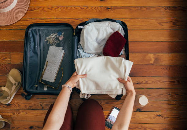 A Caucasian woman packing a suitcase from above on the wooden floor on a holiday or a business trip A Caucasian woman packing a suitcase from above on the wooden floor on a holiday or a business trip with a water bottle, a smartphone, sandals and a hat on the floor, and essentials in the spinner luggage hand luggage stock pictures, royalty-free photos & images