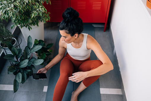 Young beautiful fit Caucasian woman sitting on a yoga mat while checking her smartphone (Horizontal, from above shot)