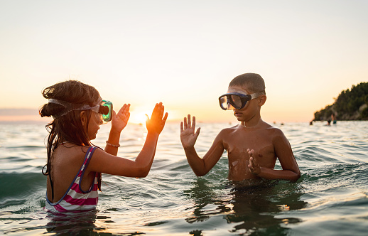 Excited and happy, boy and girl, playing a clapping game in the sea, during sunset while both wearing swimming goggles