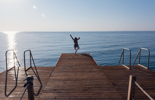 Boy jumping from wooden pier into the sea on a sunny summer day. Sunlight is shining on the sea. Summer vacation, sea, sun, childhood and fun concept.