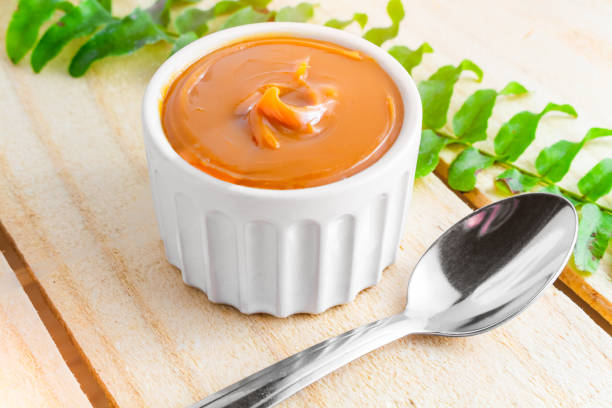 Dulce de leche in bowl on wooden farm table, decorated with branch of leaves. Image with a bowl of dulce de leche on the farm table, with a bunch of leaves and a spoon beside it lit by the morning sun. Typical sweet from France, Brazil and Argentina. dulce de leche stock pictures, royalty-free photos & images