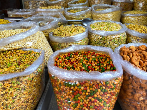 Stock photo showing a close-up view of Indian savoury salted snacks being sold by a market trader from outdoor stall display. The produce is known as Bombay mix, chevdo or namkeen. imagen de primer plano de la exhibición del puesto del mercado de namkeen (mezcla de bombay), grandes sacos de plástico de bocadillos salados salados de la india, incluidos garbanzos secos picantes, lentejas fritas, guisantes y cacahuetes, cordón, enfoq