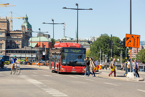 Stockholm, Sweden - July 06, 2017. Traffic on urban road in capital of Sweden. Red public bus, cars and bicycles are waiting for green signal of traffic light. People cross road at pedestrian crossing.