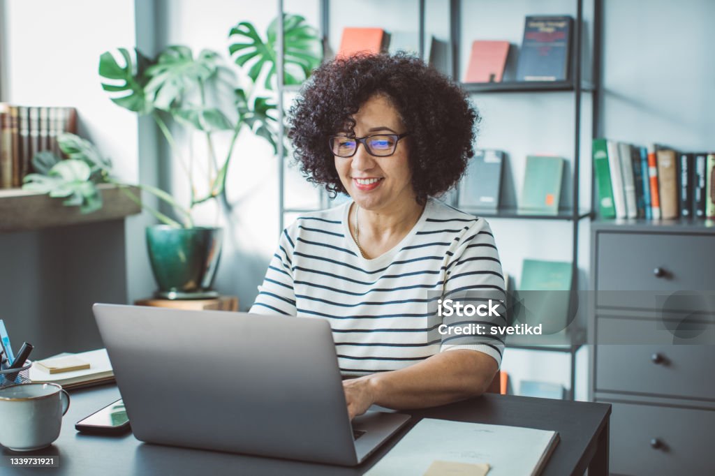 Book publisher at office Mature woman at her office. She is book publisher and reading book script or writing mail on laptop. Computer Stock Photo
