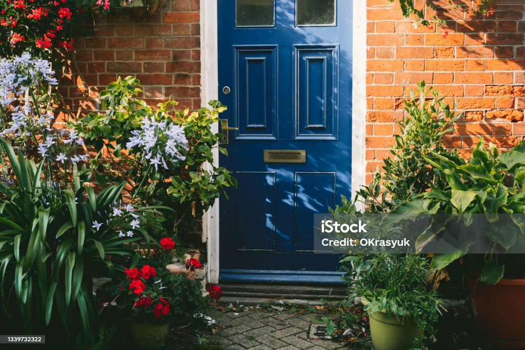 Blue front door and brick walls facade of a Beautiful Cottage with many various potted flowers. The Small garden in front of the house, gardening hobby, create a cozy space around Blue front door and brick walls facade of a Beautiful Cottage with many various potted flowers. The Small garden in front of the house, gardening hobby, create a cozy space around. Cottage Stock Photo