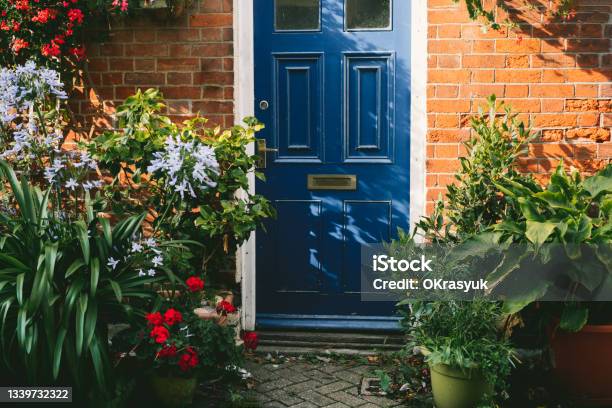 Blue Front Door And Brick Walls Facade Of A Beautiful Cottage With Many Various Potted Flowers The Small Garden In Front Of The House Gardening Hobby Create A Cozy Space Around Stock Photo - Download Image Now