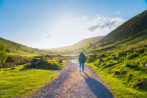 Photo of Trekking in mountains. Tourist with a backpack going on a mountain road with green hills around. Hiking alone. Sunset light. Wales, Snowdonia.