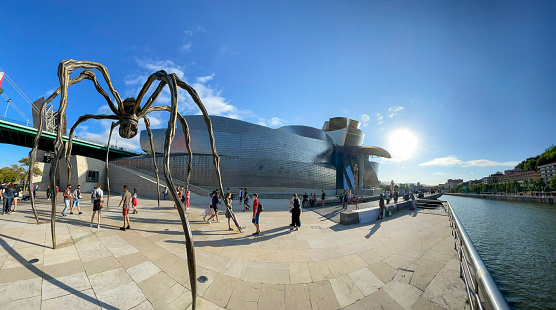 08/08/2021, Bilbao, Basque Country, Spain. Exterior view of the architecture of the Guggenheim Museum. People walk around and look at the exterior of the museum. The statue of Mom, in the foreground, which is the spider, made by Louise Bourgeois.