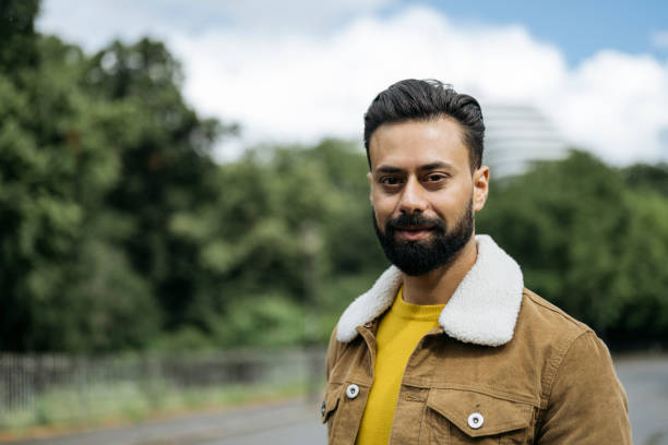 Outdoor portrait of Indian man with beard Three-quarter front view of mid adult man in gold colored sweater and tan jacket with shearling collar looking at camera with tree area in background. corduroy jacket stock pictures, royalty-free photos & images