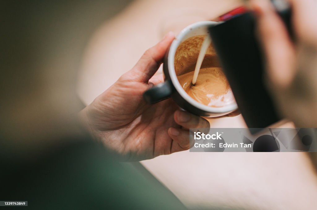 over the shoulder asian chinese male barista pouring froth milk on coffee cup prepared coffee latte art at bar counter Coffee - Drink Stock Photo