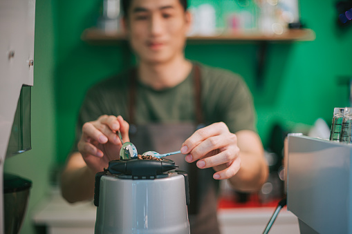 asian chinese male barista scooping filling coffee grinder with roasted coffee bean preparing for coffee grinding expresso making at cafe coffee shop