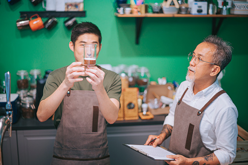 Asian Chinese senior male barista business owner training guiding new employee making coffee at bar counter of his cafe