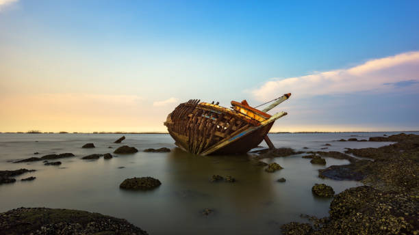 le bateau se détériore en panne couché du côté de la côte avec le coucher du soleil en arrière-plan - horizon over water white green blue photos et images de collection