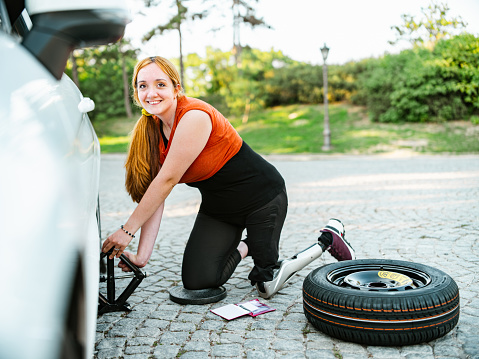 Young Woman, Leg Amputee having flat tire on the car. Young Caucasian woman, dressed in casual summer clothes with long red hair. Exterior of countryside, small white car on the side of the road.