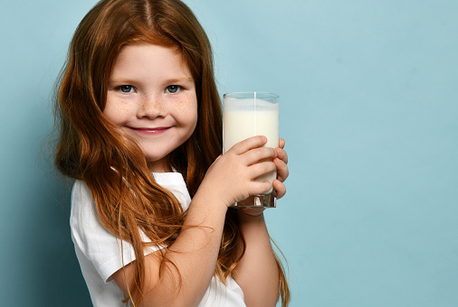 Cute red hair girl child kid drinking milk or kefir yogurt joyfully smiling on light blue background. Health and diet concept. Healthy drink breakfast and clean food concept, copy space