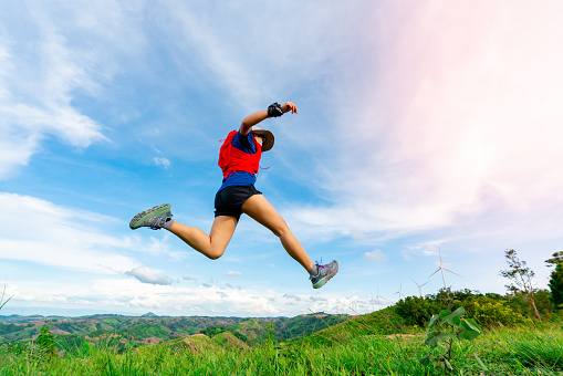 Asian female trail runners, wearing sportswear, are practicing running and jumping on the high mountain behind a beautiful view. There is a field of wind turbines generating electricity in the background.