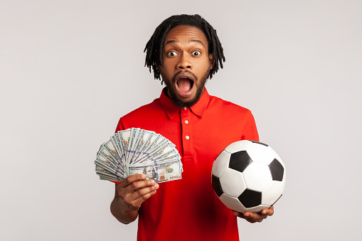 Yelling man with dreadlocks wearing red casual style T-shirt, holding soccer ball and hundred dollar banknotes, looking camera, betting and winning. Indoor studio shot isolated on gray background.