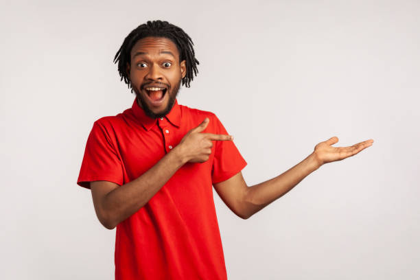 istock Handsome man with dreadlocks wearing red T-shirt, presenting copy space on his palm and looking surprised, showing empty place for promotional text. 1339797440