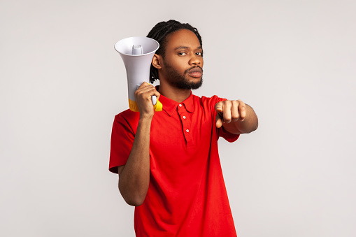 Strict serious man with dreadlocks wearing red casual style T-shirt, pointing finger at camera holding loudspeaker in hand, talking to you, protesting. Indoor studio shot isolated on gray background.
