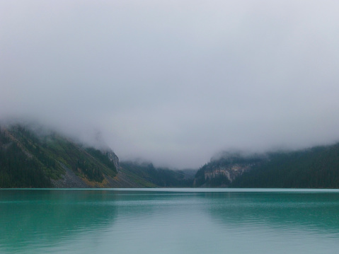 Green-blue Canadian lake in front of misty mountain with green conifer trees
