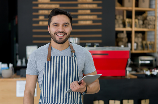 Portrait of a happy Latin American waiter working at a cafe using a tablet computer and looking at the camera smiling- food service concepts