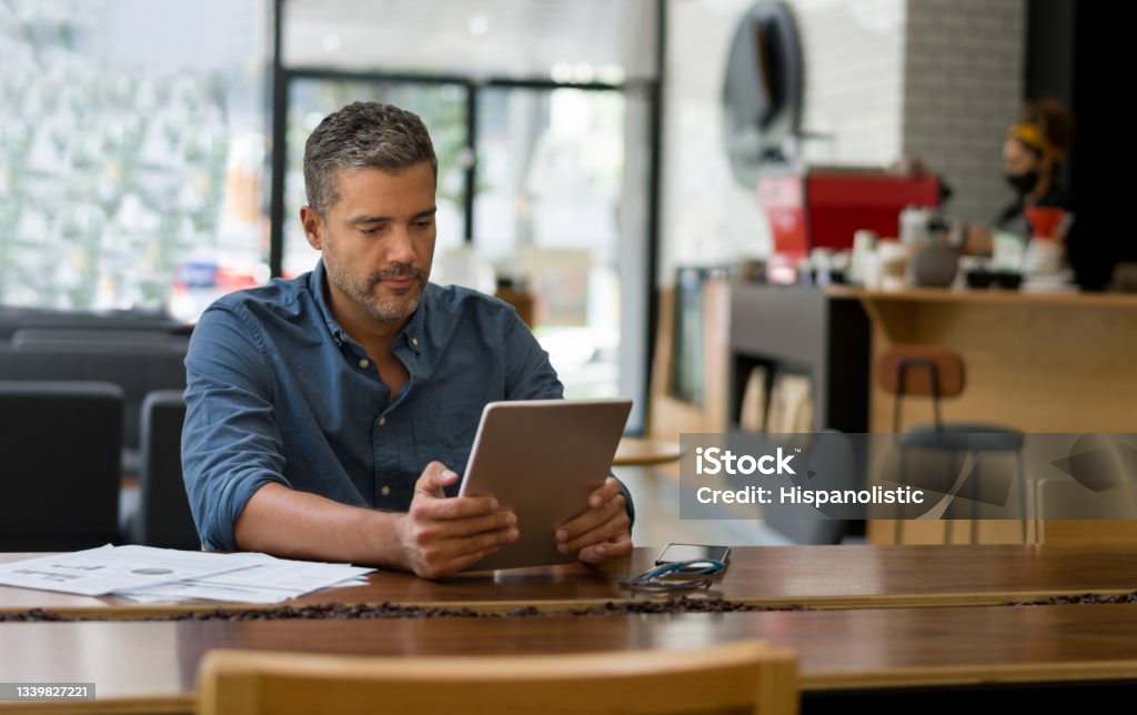 Business man working at a cafe on his tablet while waiting for a coffee Happy Latin American business man working at a cafe on his tablet while waiting for a coffee - lifestyle concepts. **DOCUMENTS WERE MADE FROM SCRATCH BY US** Men Stock Photo