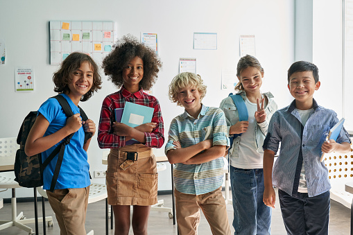 Portrait of cheerful smiling diverse schoolchildren standing posing in classroom holding notebooks and backpacks looking at camera happy after school reopen. Diversity. Back to school concept.