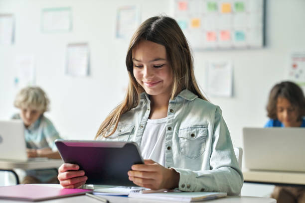 Happy smiling schoolgirl reading task from tablet computer device in classroom.