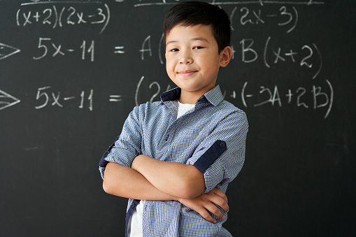 Happy cheerful proud confident smiling cute asian schoolboy with arms crossed standing posing in classroom on chalkboard blackboard with formulas background. Elementary preteen school kid portrait.