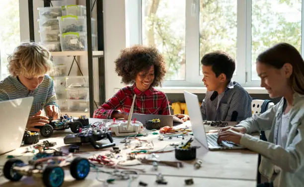 Group of multiethnic schoolkids interacting using gadgets for programming for robotics engineering class. Elementary school science classroom of futuristic technologies. STEM education concept.