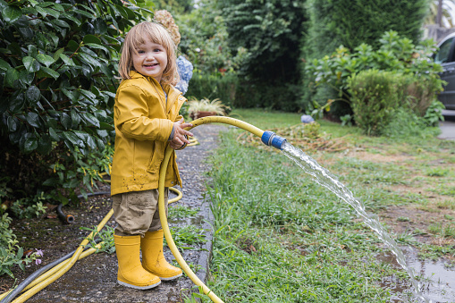 Side view of cute little child wearing yellow raincoat and rubber boots watering garden from hose after rain and looking at camera. Concept children helping with housework